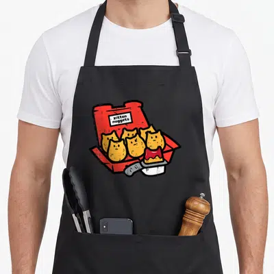 Nugget kitten apron - Six yellow kittens in red fry box labeled Kitten nuggets with sauce container — shown on a black cotton canvas apron against a white background