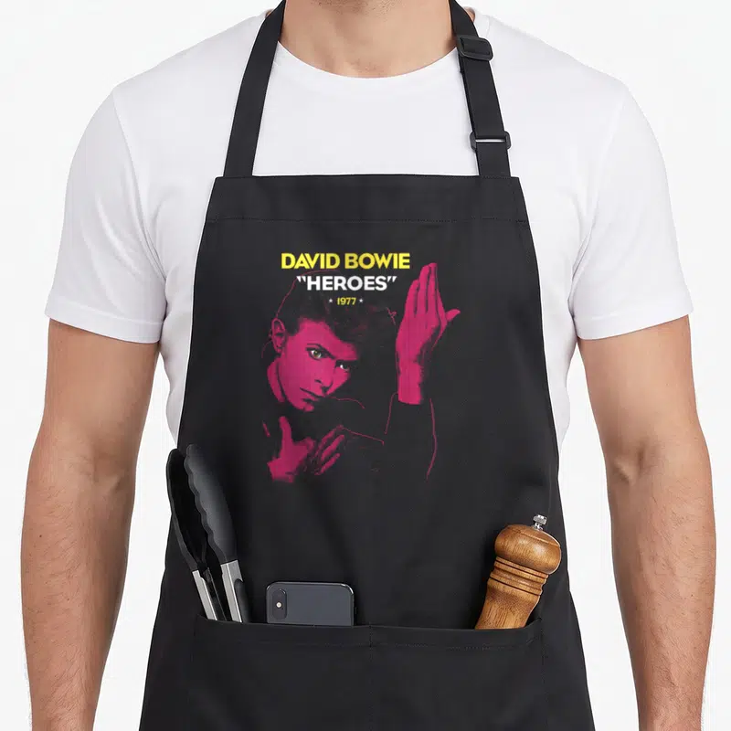Heroe david aprons - Pink David Bowie portrait with raised hand and yellow 1977 Heroes text — shown on a black cotton canvas apron against a white background
