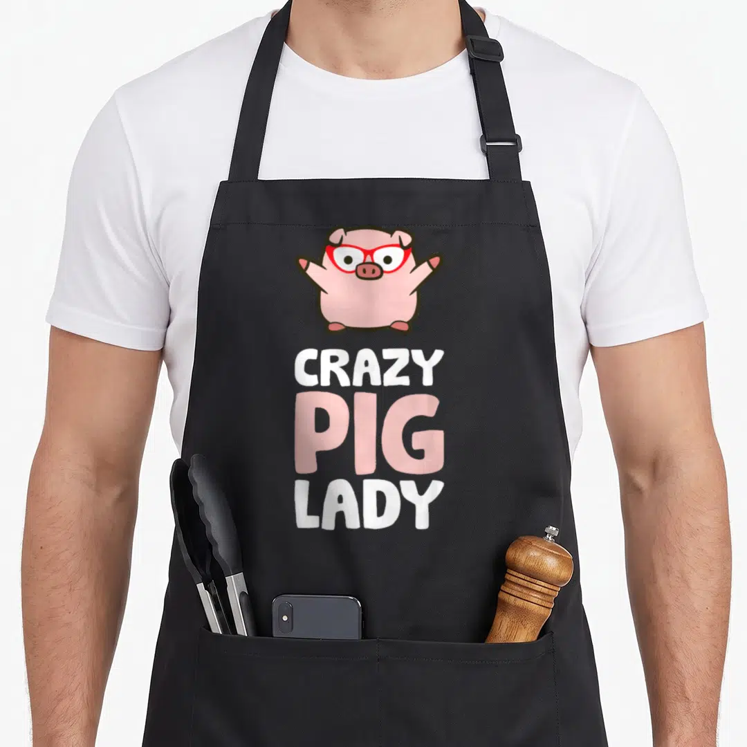 Retro crazy pig man apron - Cartoon pink pig with red glasses above stacked white and pink text reading CRAZY PIG LADY — shown on a black cotton canvas apron against a white background