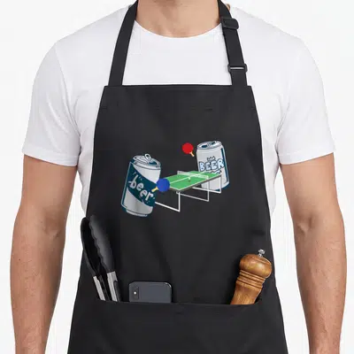 Playing beer pong apron - Two cartoon beer cans playing ping pong on a green table with red and blue paddles — shown on a black cotton canvas apron against a white background