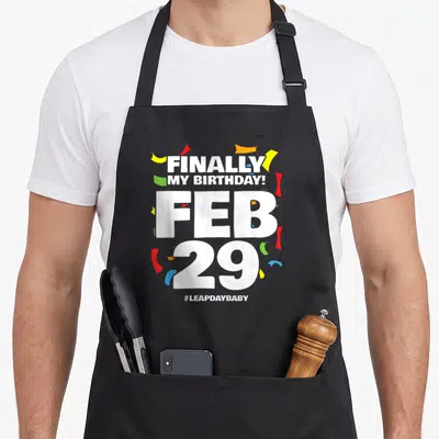 Leap baby birthday apron - Finally My Birthday Feb 29 text with colorful confetti and hashtag leapdaybaby — shown on a black cotton canvas apron against a white background