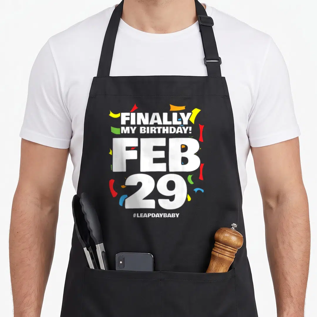 Leap baby birthday apron - Finally My Birthday Feb 29 text with colorful confetti and hashtag leapdaybaby — shown on a black cotton canvas apron against a white background