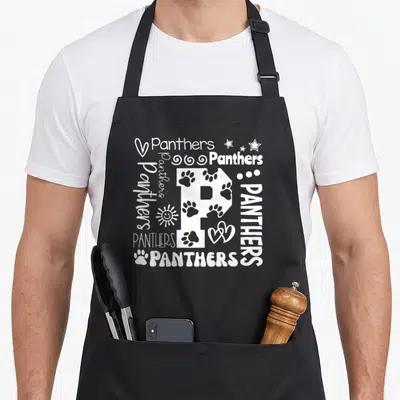School novelty apron - White letter P surrounded by Panthers text, paw prints, stars, hearts, and sun icons — shown on a black cotton canvas apron against a white background