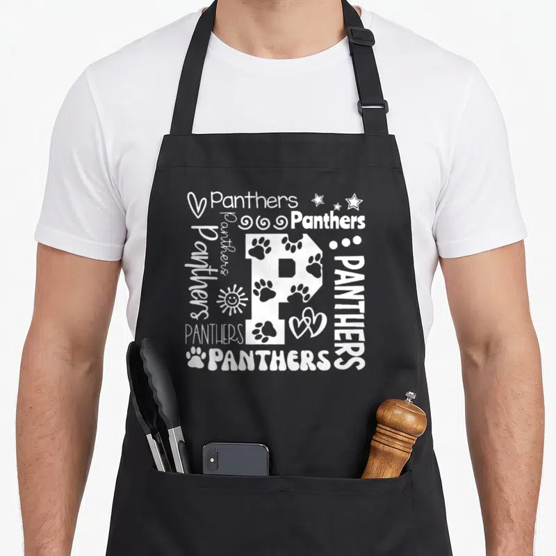School novelty apron - White letter P surrounded by Panthers text, paw prints, stars, hearts, and sun icons — shown on a black cotton canvas apron against a white background