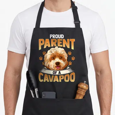 Proud parent dog apron - Cartoon Cavapoo head with orange text and scattered paw prints — shown on a black cotton canvas apron against a white background