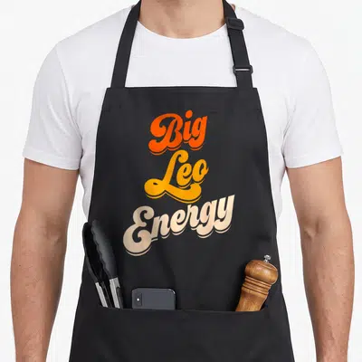 Energy leo apron - Stacked Big Leo Energy text in orange yellow and cream script — shown on a black cotton canvas apron against a white background