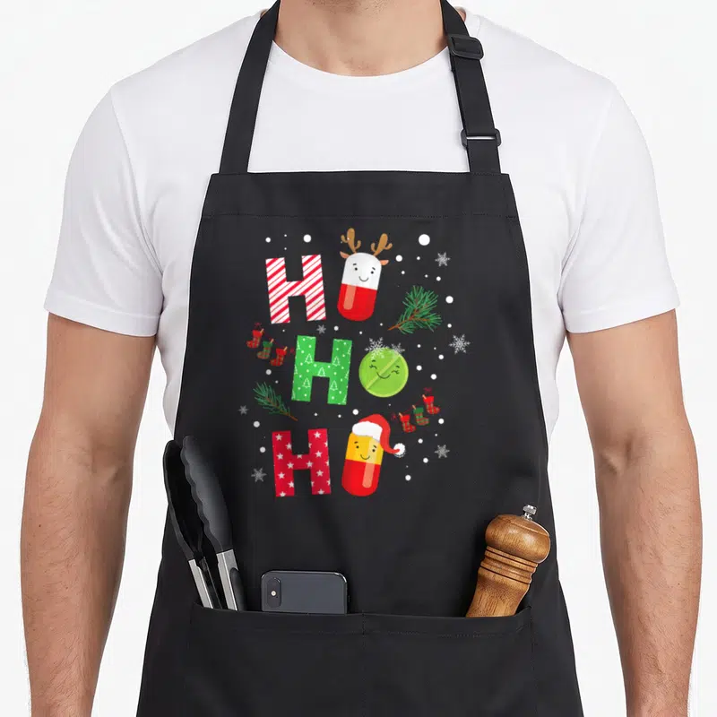 Christmas pharmacist apron - Stacked HO HO HO text with cartoon pills, pine branches, stockings, and snowflakes in red — shown on a black cotton canvas apron against a white background