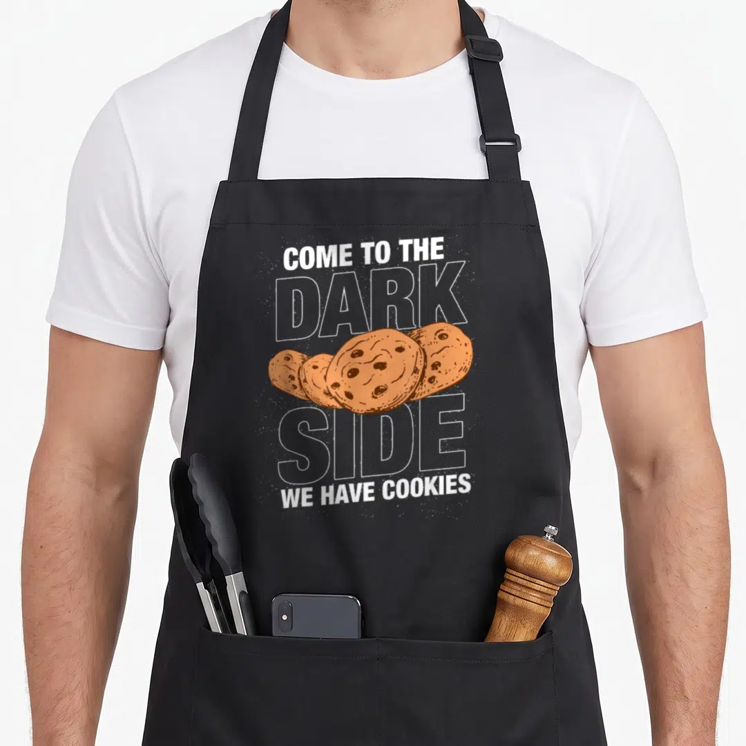 Come cookies apron - Stacked text reading come to the dark side we have cookies above and below overlapping brown — shown on a black cotton canvas apron against a white background