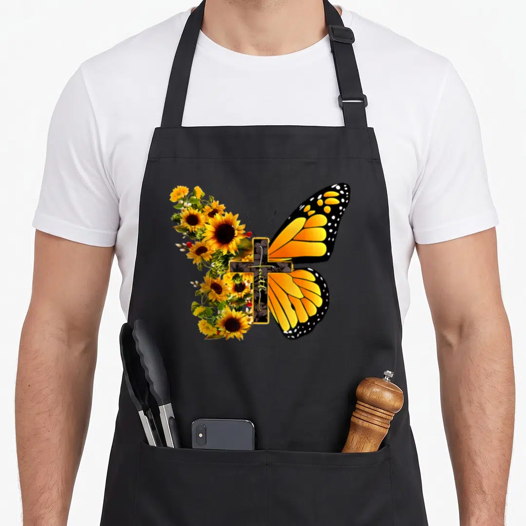 Butterfly funny christian aprons - Split butterfly with sunflower left wing and orange right wing, cross with vertical — shown on a black cotton canvas apron against a white background