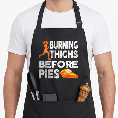 Running pie apron - Orange runner silhouette and white distressed text burning thighs before pies next to a pie slice — shown on a black cotton canvas apron against a white background
