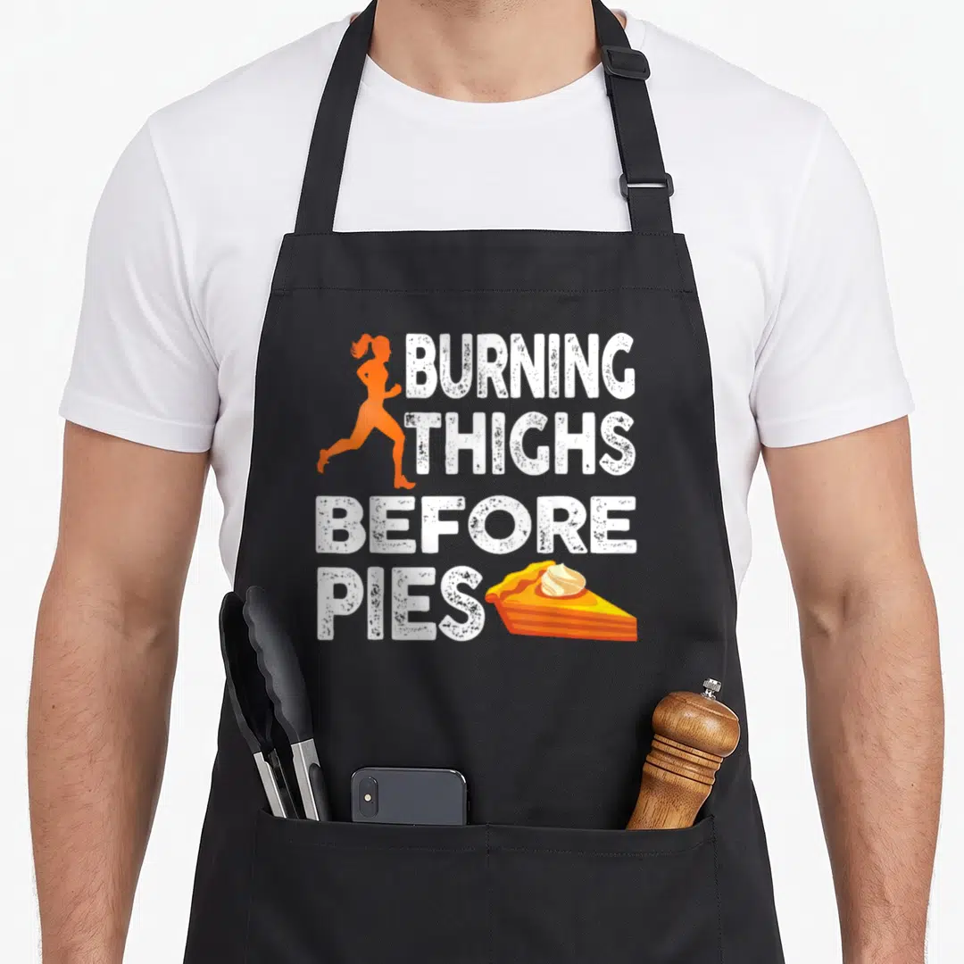 Running pie apron - Orange runner silhouette and white distressed text burning thighs before pies next to a pie slice — shown on a black cotton canvas apron against a white background