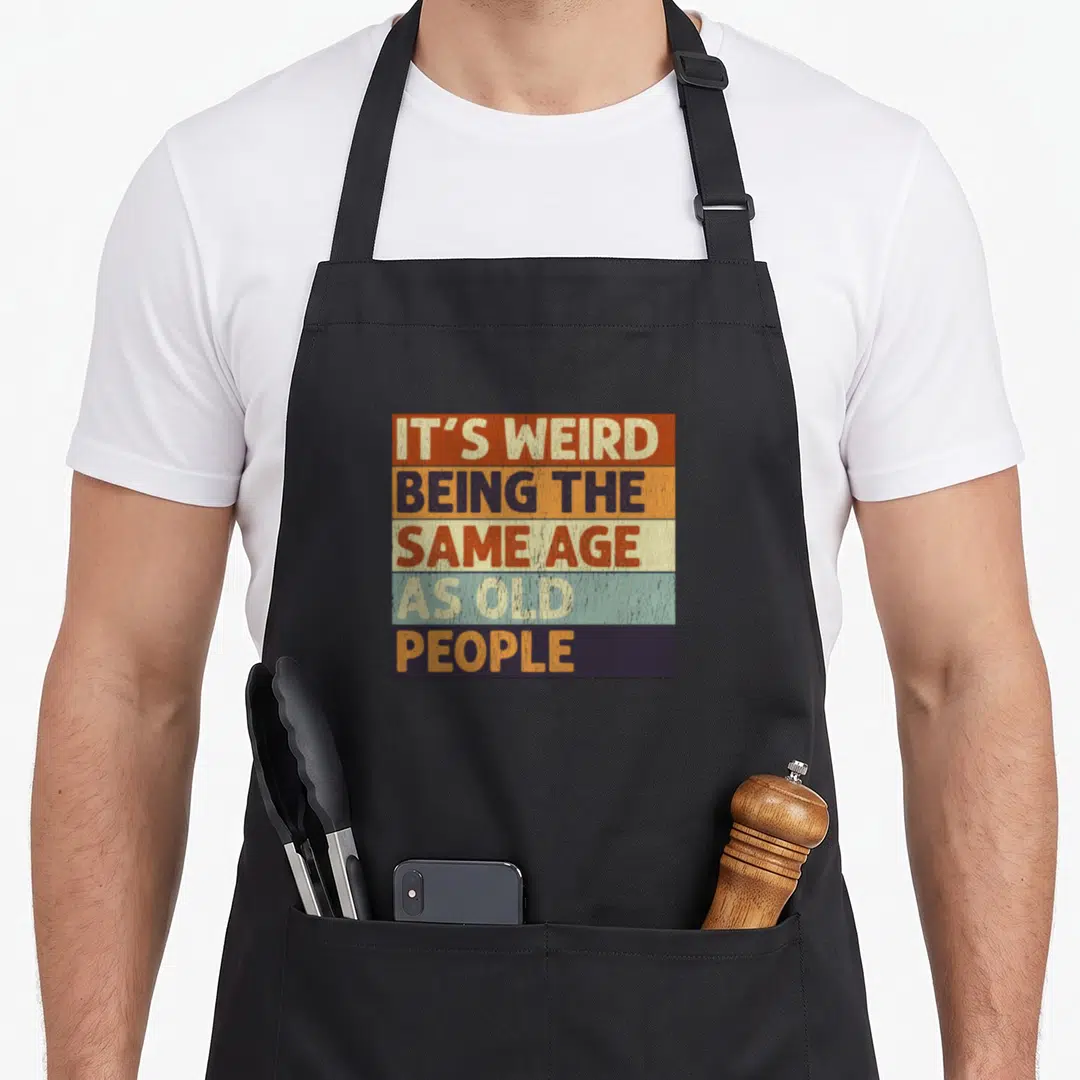 Vintage sarcastic weird aprons - Retro text IT'S WEIRD BEING THE SAME AGE AS OLD PEOPLE on orange blue beige green — shown on a black cotton canvas apron against a white background