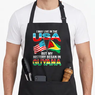 Proud guyana apron - Crossed US and Guyana flags with text I MAY LIVE IN THE USA BUT MY HISTORY BEGAN IN GUYANA — shown on a black cotton canvas apron against a white background