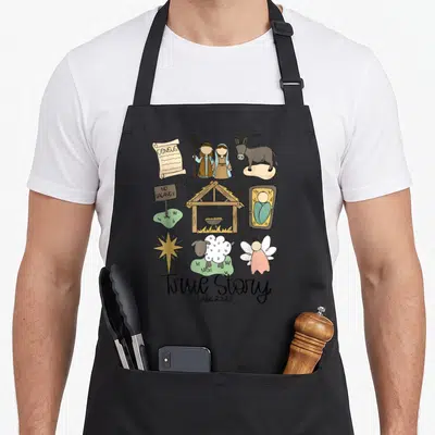 Nativity christian christmas apron - Cartoon nativity icons with census scroll, no vacancy sign, and True Story Luke — shown on a black cotton canvas apron against a white background