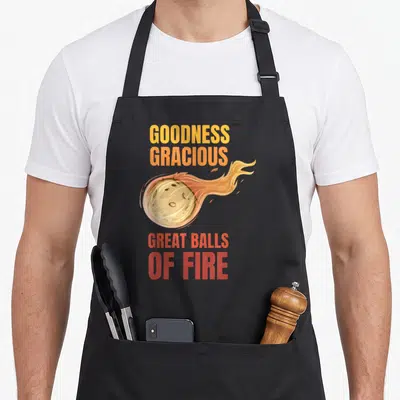 Ball asteroid apron - Cartoon meteor with fire trail and stacked text GOODNESS GRACIOUS GREAT BALLS OF FIRE in yellow — shown on a black cotton canvas apron against a white background