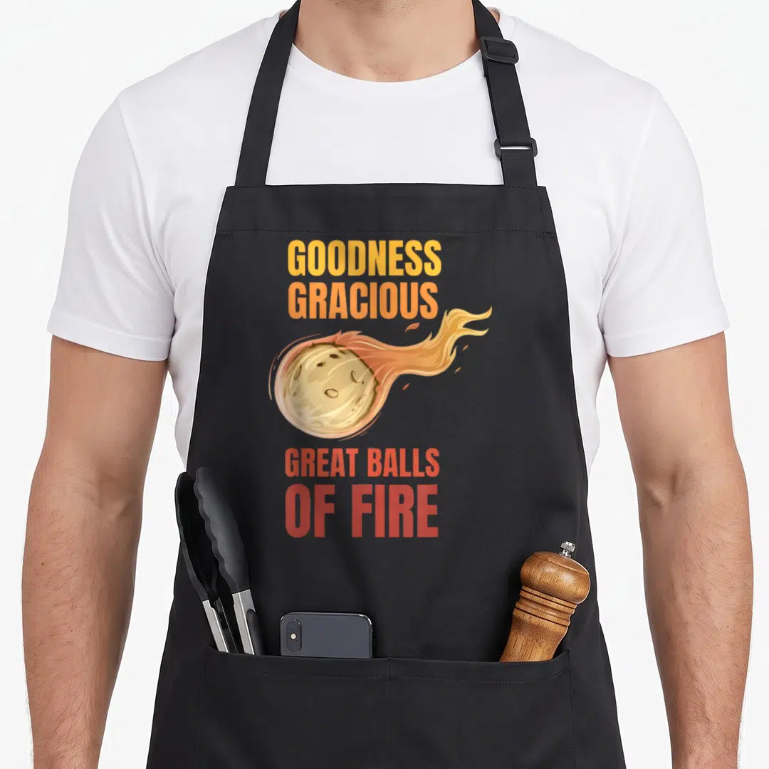 Ball asteroid apron - Cartoon meteor with fire trail and stacked text GOODNESS GRACIOUS GREAT BALLS OF FIRE in yellow — shown on a black cotton canvas apron against a white background