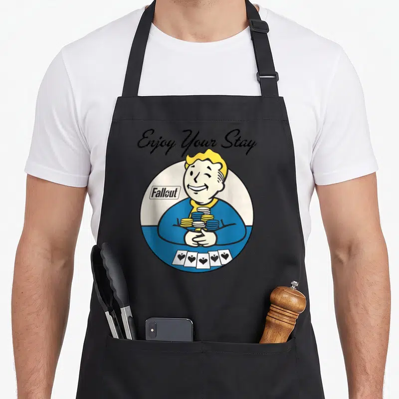 Vault video game apron - Vault Boy winking holding gold and blue coins with five playing cards below arched text — shown on a black cotton canvas apron against a white background