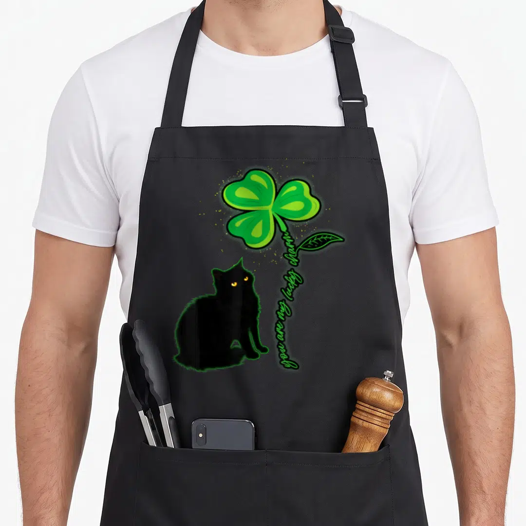 Patrick black cat apron - Black cat sitting beside green four-leaf clover with vertical lucky charm text — shown on a black cotton canvas apron against a white background