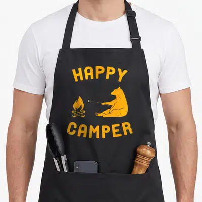 Bonfire happy camper apron - Orange cartoon bear roasting marshmallow with HAPPY CAMPER text arched above and below — shown on a black cotton canvas apron against a white background