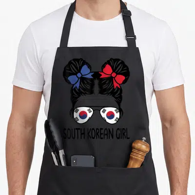 South messy aprons - Cartoon girl with space buns and South Korean flag sunglasses. Text reads SOUTH KOREAN GIRL below — shown on a black cotton canvas apron against a white background