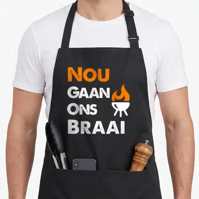 Nou south african apron - Orange and white Nou Gaan Ons Braai text with grill icon on distressed background — shown on a black cotton canvas apron against a white background