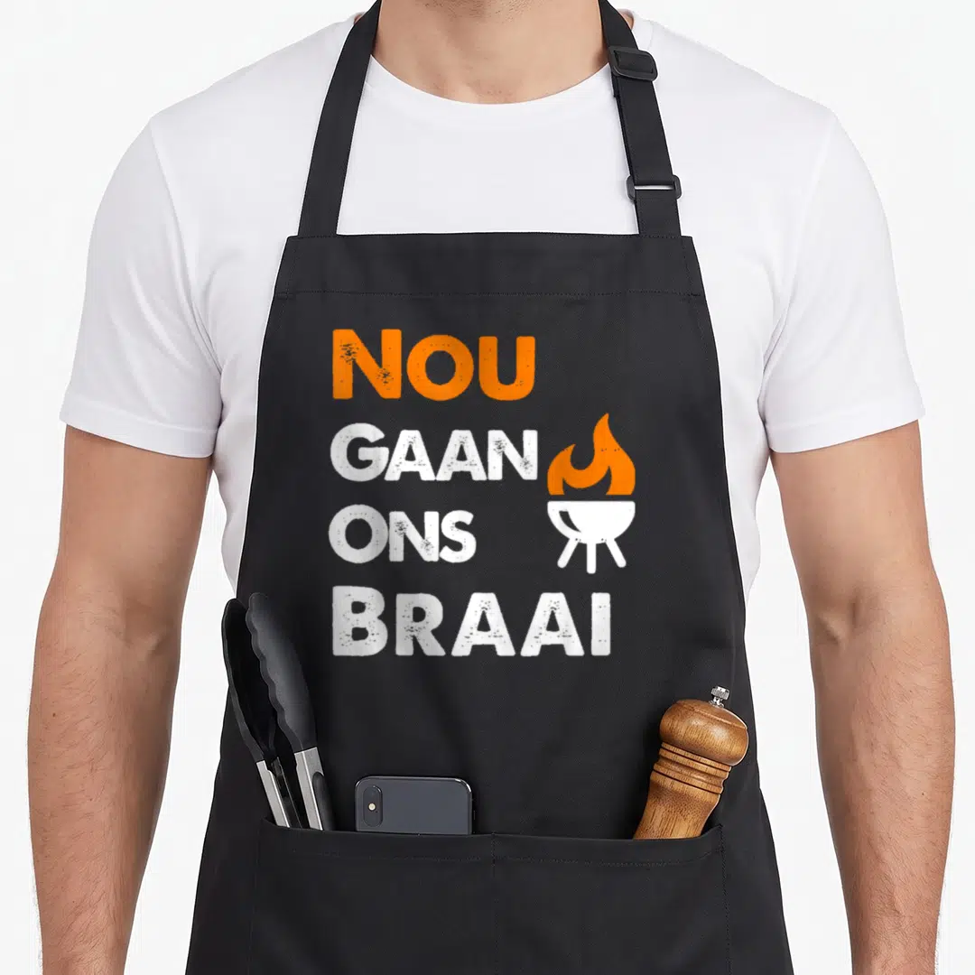 Nou south african apron - Orange and white Nou Gaan Ons Braai text with grill icon on distressed background — shown on a black cotton canvas apron against a white background