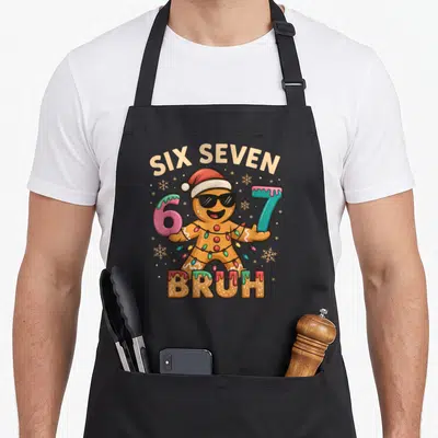 Six christmas apron - Cartoon gingerbread man with sunglasses holding pink 6 and blue 7 under six seven text — shown on a black cotton canvas apron against a white background