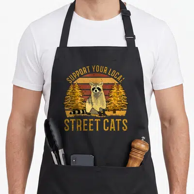 Support local cat apron - Raccoon standing between two pine trees in front of a striped retro sun with text — shown on a black cotton canvas apron against a white background