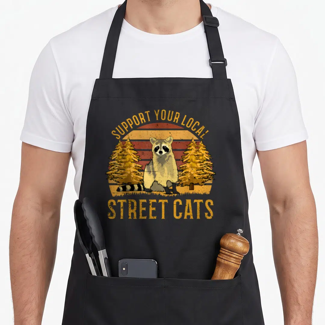 Support local cat apron - Raccoon standing between two pine trees in front of a striped retro sun with text — shown on a black cotton canvas apron against a white background