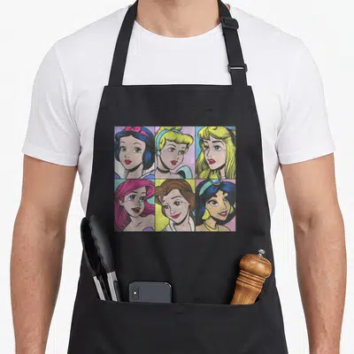 Pop princess apron - Six cartoon princess portraits in colored panels arranged in a two by three grid — shown on a black cotton canvas apron against a white background