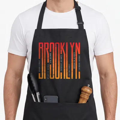Brooklyn illustration new york apron - Vertical BROOKLYN text filled with street names like Flatbush Avenue and Grand — shown on a black cotton canvas apron against a white background