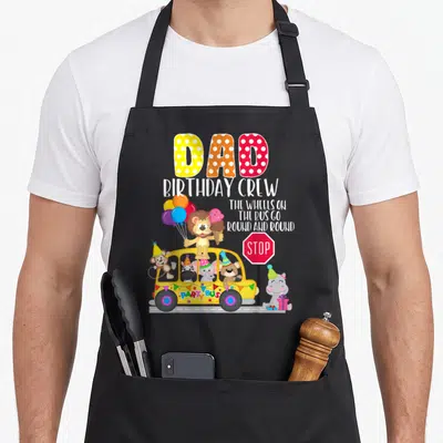 Bus birthday apron - DAD BIRTHDAY CREW text above yellow party bus with lion, hippo, and zoo animals holding balloons — shown on a black cotton canvas apron against a white background