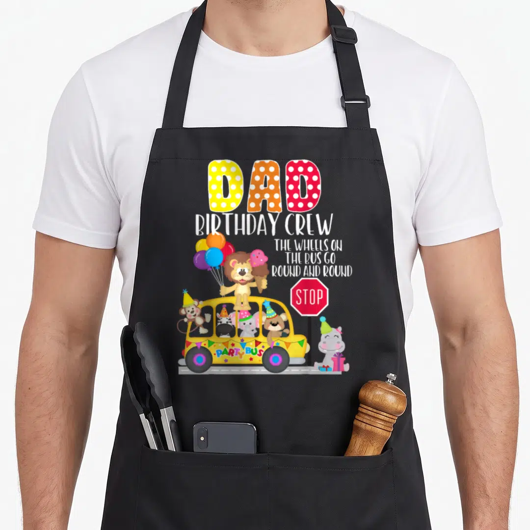 Bus birthday apron - DAD BIRTHDAY CREW text above yellow party bus with lion, hippo, and zoo animals holding balloons — shown on a black cotton canvas apron against a white background