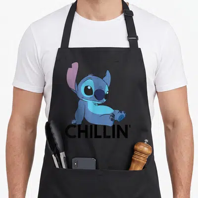 Funny chillin stitch apron - Blue cartoon character sitting above bold black CHILLIN' text — shown on a black cotton canvas apron against a white background