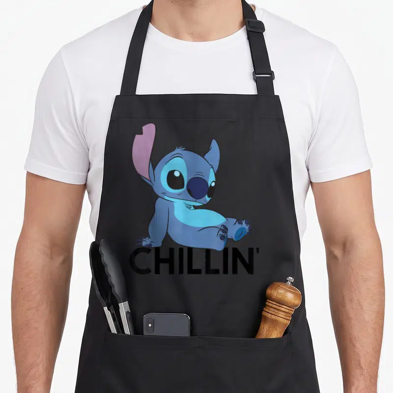 Funny chillin stitch apron - Blue cartoon character sitting above bold black CHILLIN' text — shown on a black cotton canvas apron against a white background