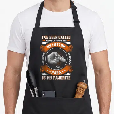 Lot papa apron - Orange and white text surrounds a grey circle showing a baby hand holding an adult finger flanked — shown on a black cotton canvas apron against a white background