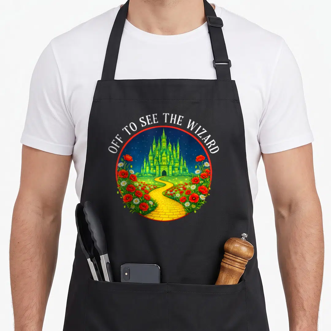 Road wicked apron - Green castle on yellow brick road with red poppies and white daisies under text Off to See — shown on a black cotton canvas apron against a white background