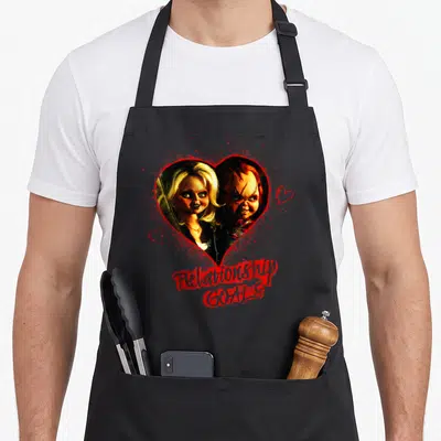 Play novelty apron - Two dolls inside red heart with Relationship GOALS text and smaller hearts — shown on a black cotton canvas apron against a white background