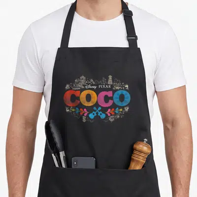 Coco colored aprons - COCO text in red yellow pink blue with two guitars and skyline — shown on a black cotton canvas apron against a white background