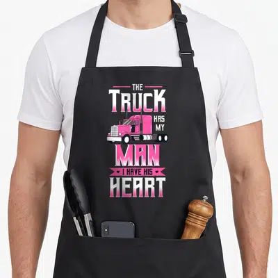 Truck driving aprons - Pink retro truck graphic with stacked text THE TRUCK HAS MY MAN I HAVE HIS HEART and two small — shown on a black cotton canvas apron against a white background