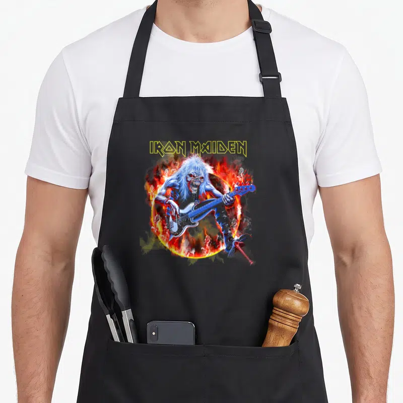 Fear flame apron - IRON MAIDEN text above blue zombie playing bass in orange and red fire ring — shown on a black cotton canvas apron against a white background
