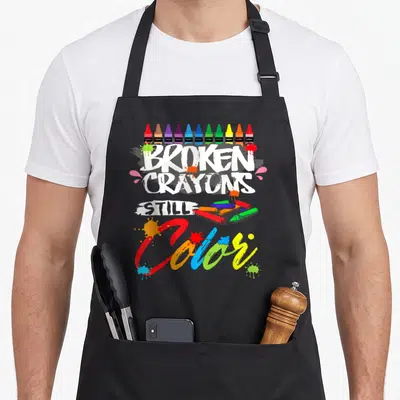 Broken crayons apron - Stacked text reading BROKEN CRAYONS STILL Color with crayon illustrations — shown on a black cotton canvas apron against a white background
