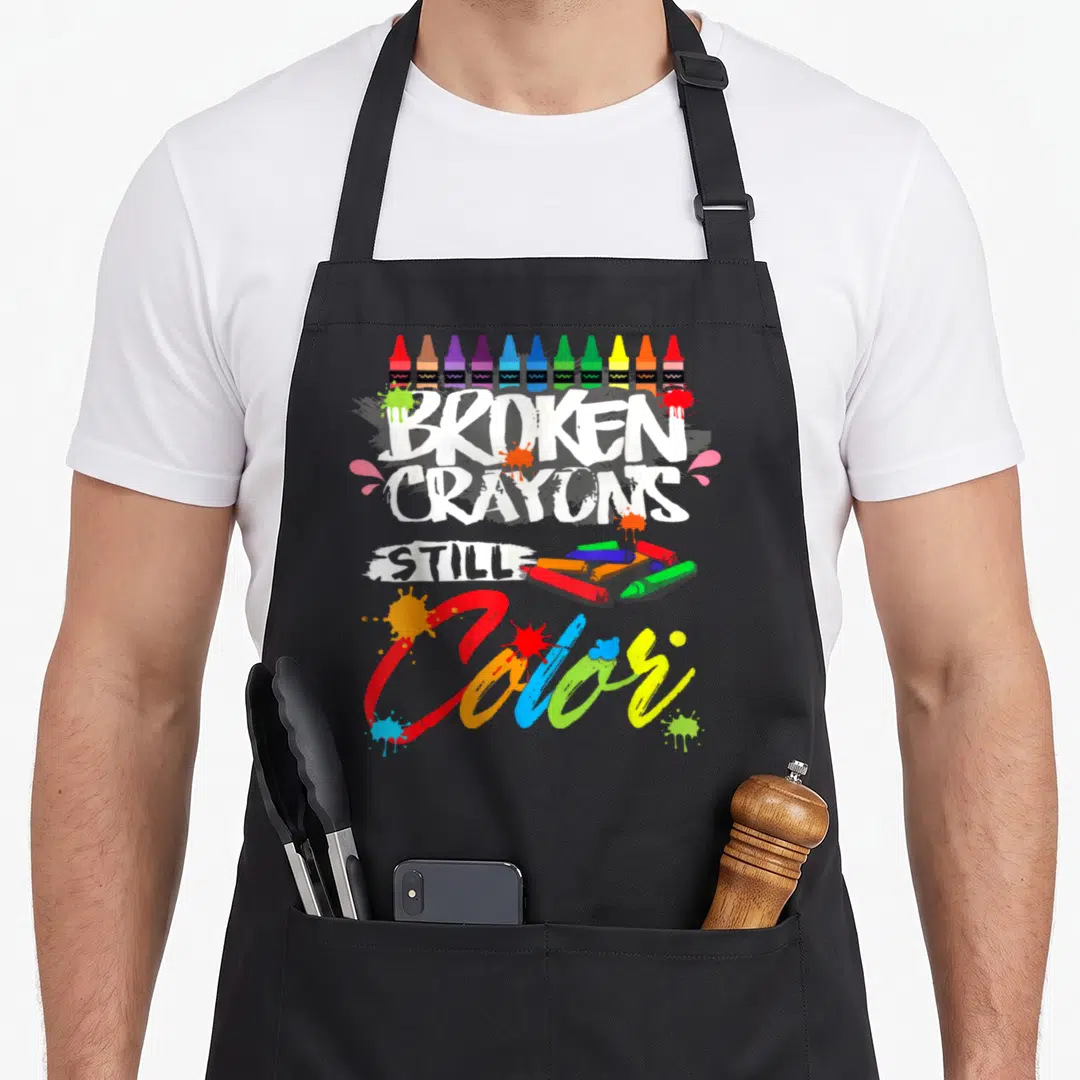 Broken crayons apron - Stacked text reading BROKEN CRAYONS STILL Color with crayon illustrations — shown on a black cotton canvas apron against a white background