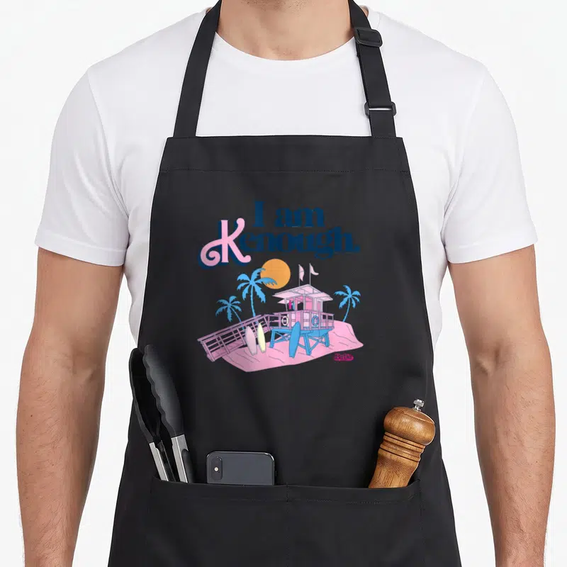 Lifeguard i am kenough apron - Cartoon beach hut with surfboards and I am Kenough text in pink and blue — shown on a black cotton canvas apron against a white background