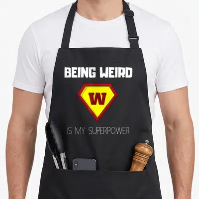 Superpower weird aprons - White text reading BEING WEIRD above a red and yellow diamond W emblem — shown on a black cotton canvas apron against a white background