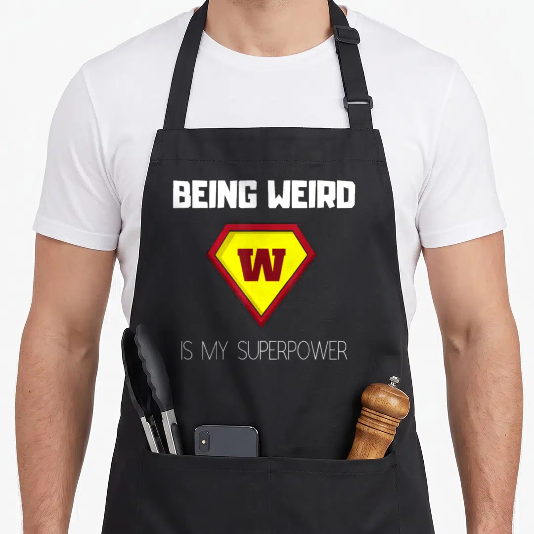 Superpower weird aprons - White text reading BEING WEIRD above a red and yellow diamond W emblem — shown on a black cotton canvas apron against a white background