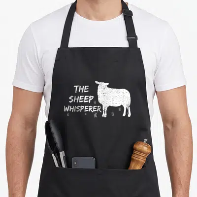 Sheep farmers apron - THE SHEEP WHISPERER text stacked left of standing white sheep with grass tufts at feet — shown on a black cotton canvas apron against a white background
