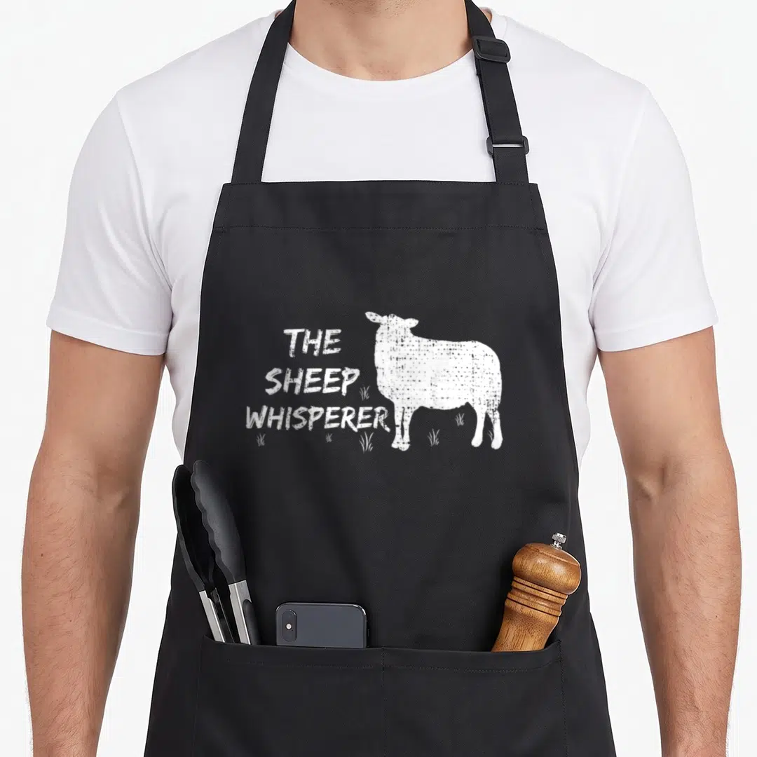 Sheep farmers apron - THE SHEEP WHISPERER text stacked left of standing white sheep with grass tufts at feet — shown on a black cotton canvas apron against a white background