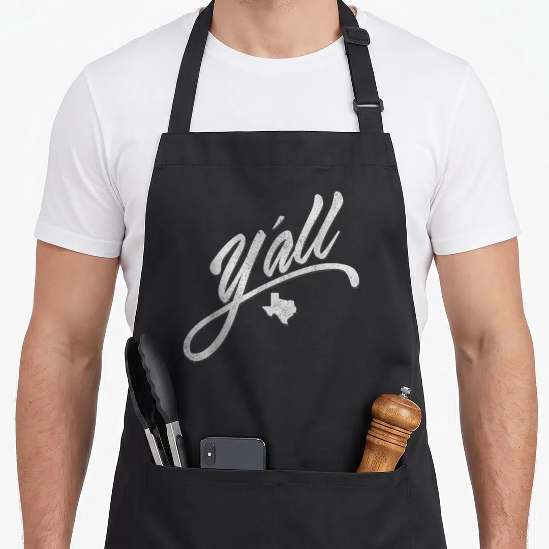 Yall texas aprons - White distressed script text y'all above a small Texas state outline on dark fabric — shown on a black cotton canvas apron against a white background