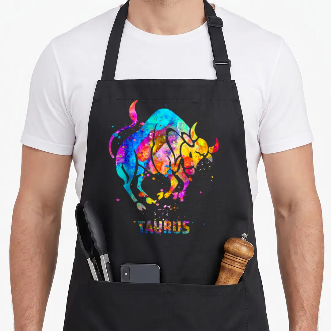 Sign taurus aprons - Rainbow watercolor bull illustration above TAURUS text in blue yellow red purple green — shown on a black cotton canvas apron against a white background